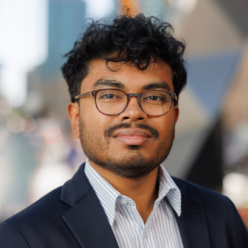 A young man with glasses wearing a navy blazer and a striped button-down shirt.