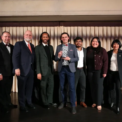 Seven people in formal attire stand in a row on a stage, with the center person holding an award.