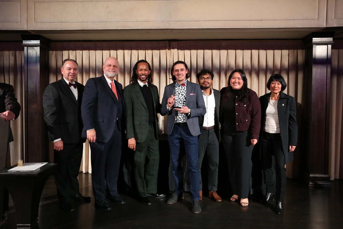 Seven people in formal attire stand in a row on a stage, with the center person holding an award.