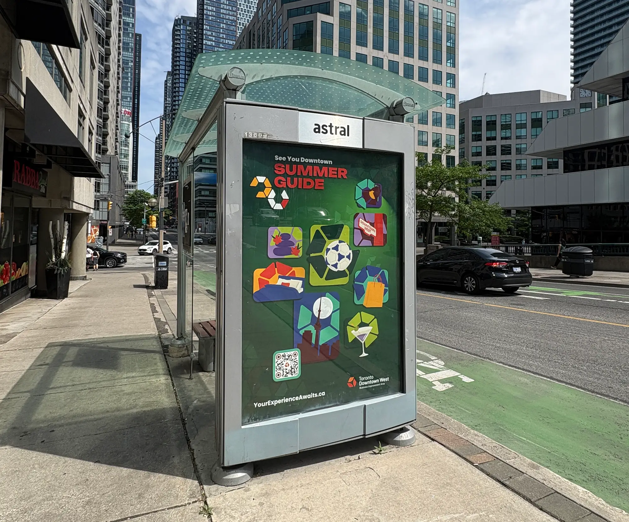 A bus shelter on a city sidewalk featuring a Summer Guide advertisement with colorful icons and a QR code.