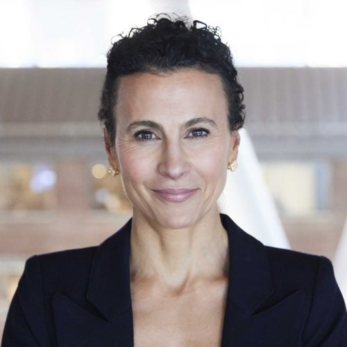 A woman with short, curly dark hair wearing a navy blazer.