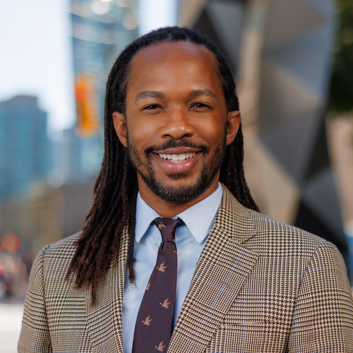 A man with dreadlocks wearing a light blue dress shirt, patterned tie, and a plaid blazer smiles at the camera.