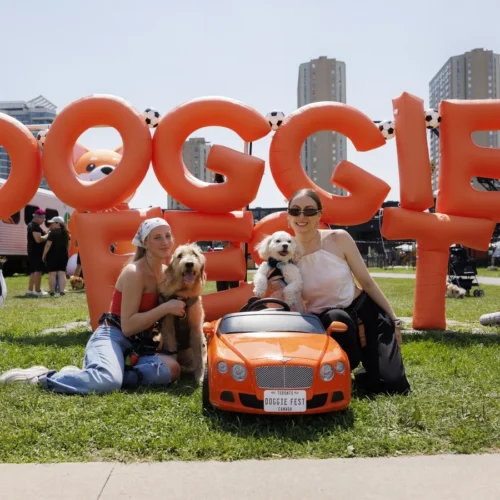 Two women with dogs sit in front of an inflatable Doggie Fest sign and a small toy car.