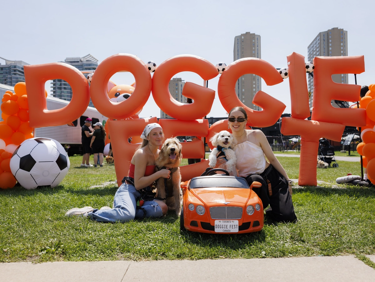 Two women with dogs sit in front of an inflatable Doggie Fest sign and a small toy car.