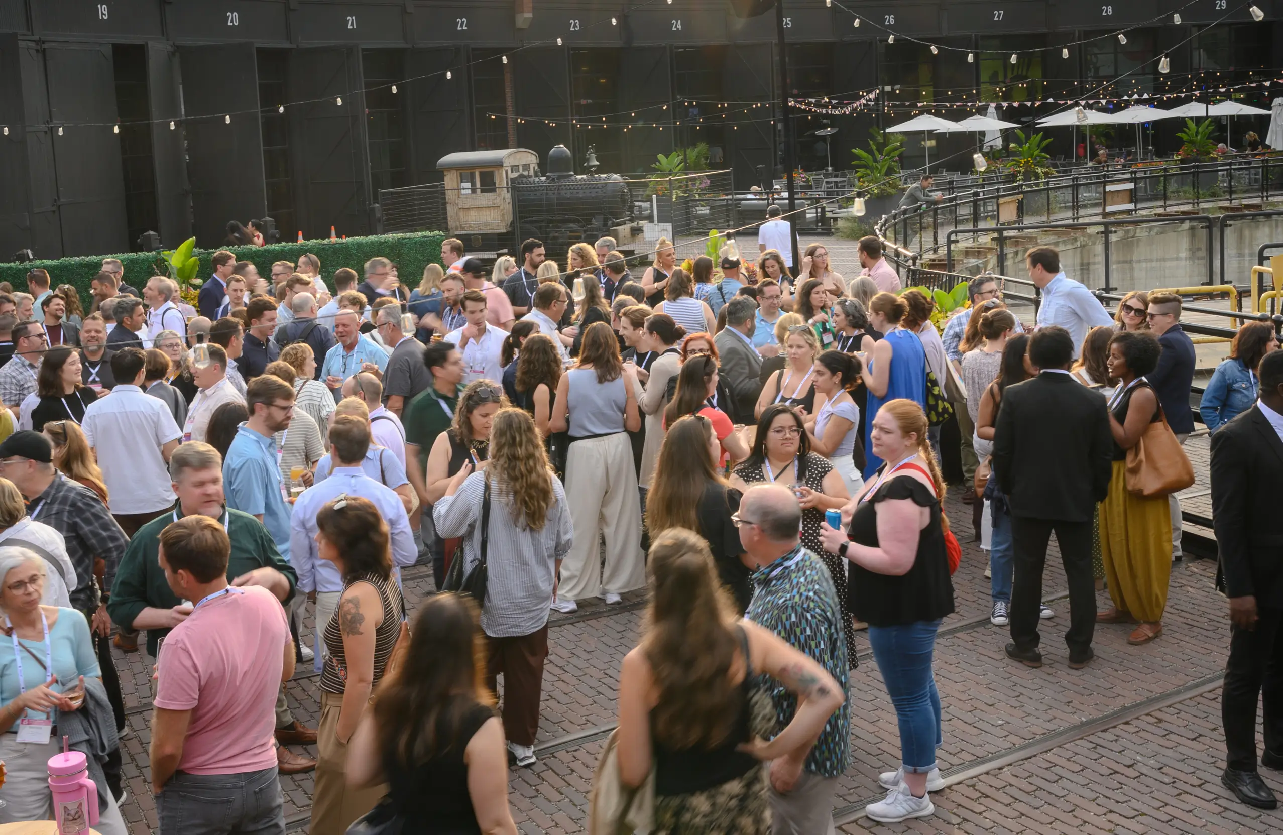 A large, diverse crowd of people mingling outdoors on a brick patio in front of a vintage train engine.