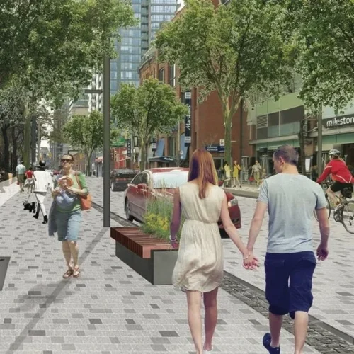 Pedestrians walk along a paved city street lined with trees, street furniture, and shops.