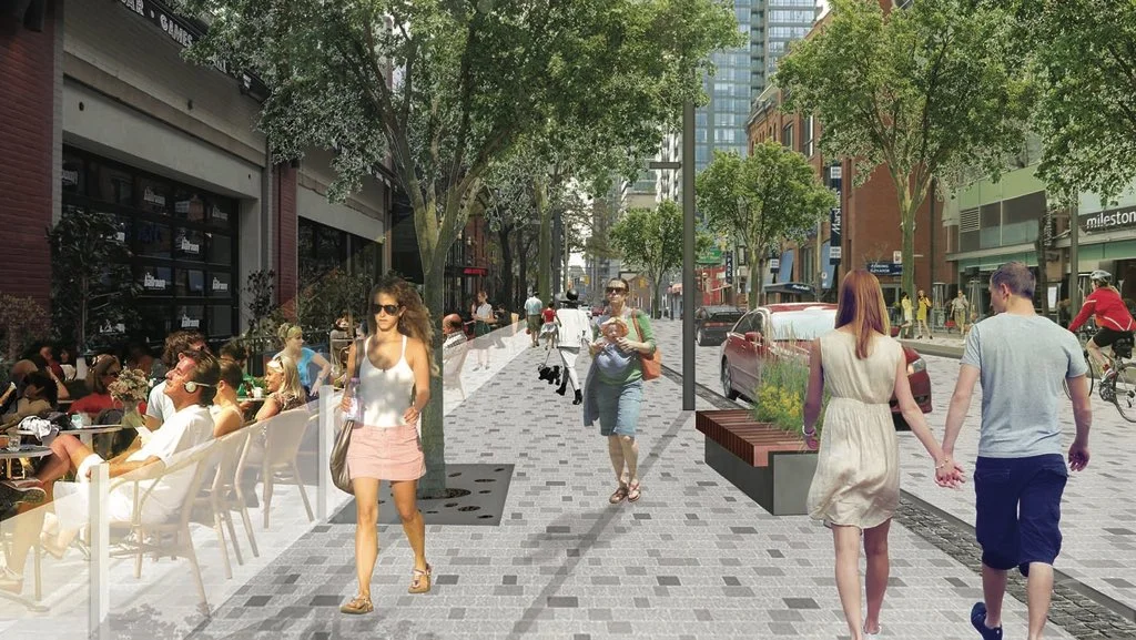 Pedestrians walk along a paved city street lined with trees, street furniture, and shops.