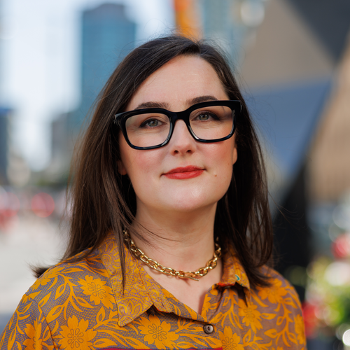 A woman with long brown hair wearing black-rimmed glasses and a yellow floral shirt.