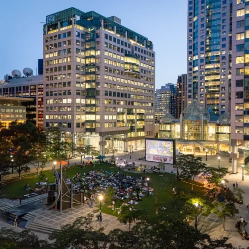 People sitting on a lawn at night watching a large outdoor movie screen in a city plaza surrounded by skyscrapers.