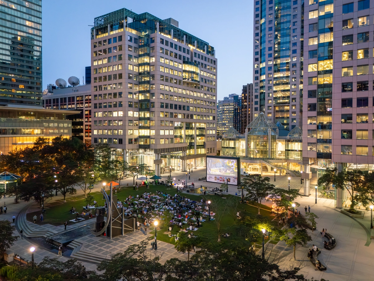 People sitting on a lawn at night watching a large outdoor movie screen in a city plaza surrounded by skyscrapers.