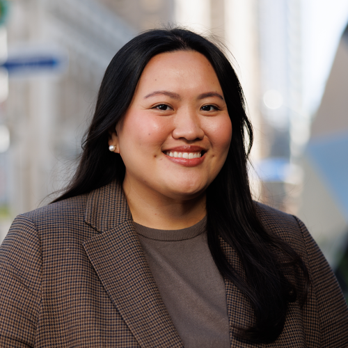 A woman with long dark hair smiles while wearing a brown blazer.