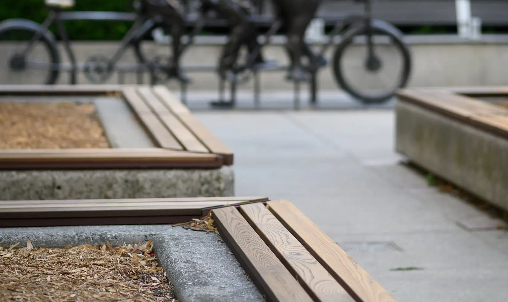Wooden benches with concrete bases in a park, with a blurred sculpture of cyclists in the background.