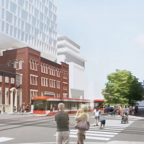 People waiting to cross a street in front of a red streetcar and a historic brick building.