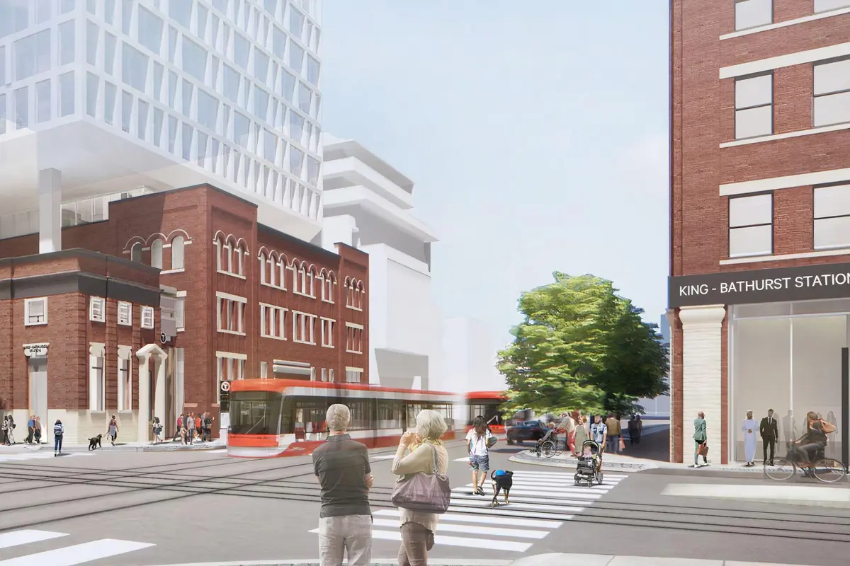 People waiting to cross a street in front of a red streetcar and a historic brick building.