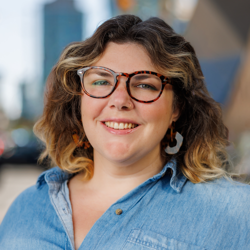 A smiling woman with shoulder-length wavy hair wears tortoiseshell glasses and a denim shirt.