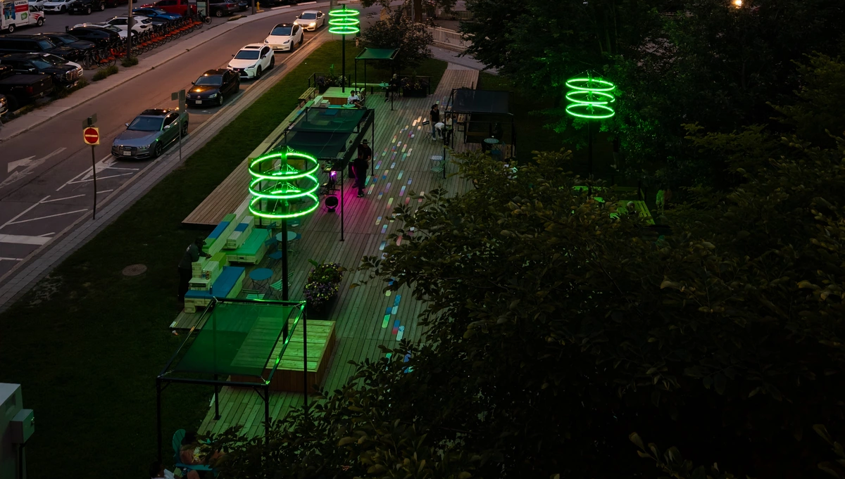 An outdoor patio at dusk featuring green neon light sculptures and wooden decking next to a street with parked cars.