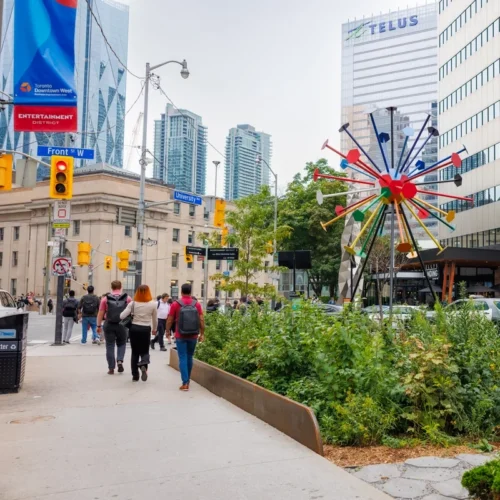 Pedestrians walk on a downtown street corner near a colorful, sunburst-shaped sculpture.