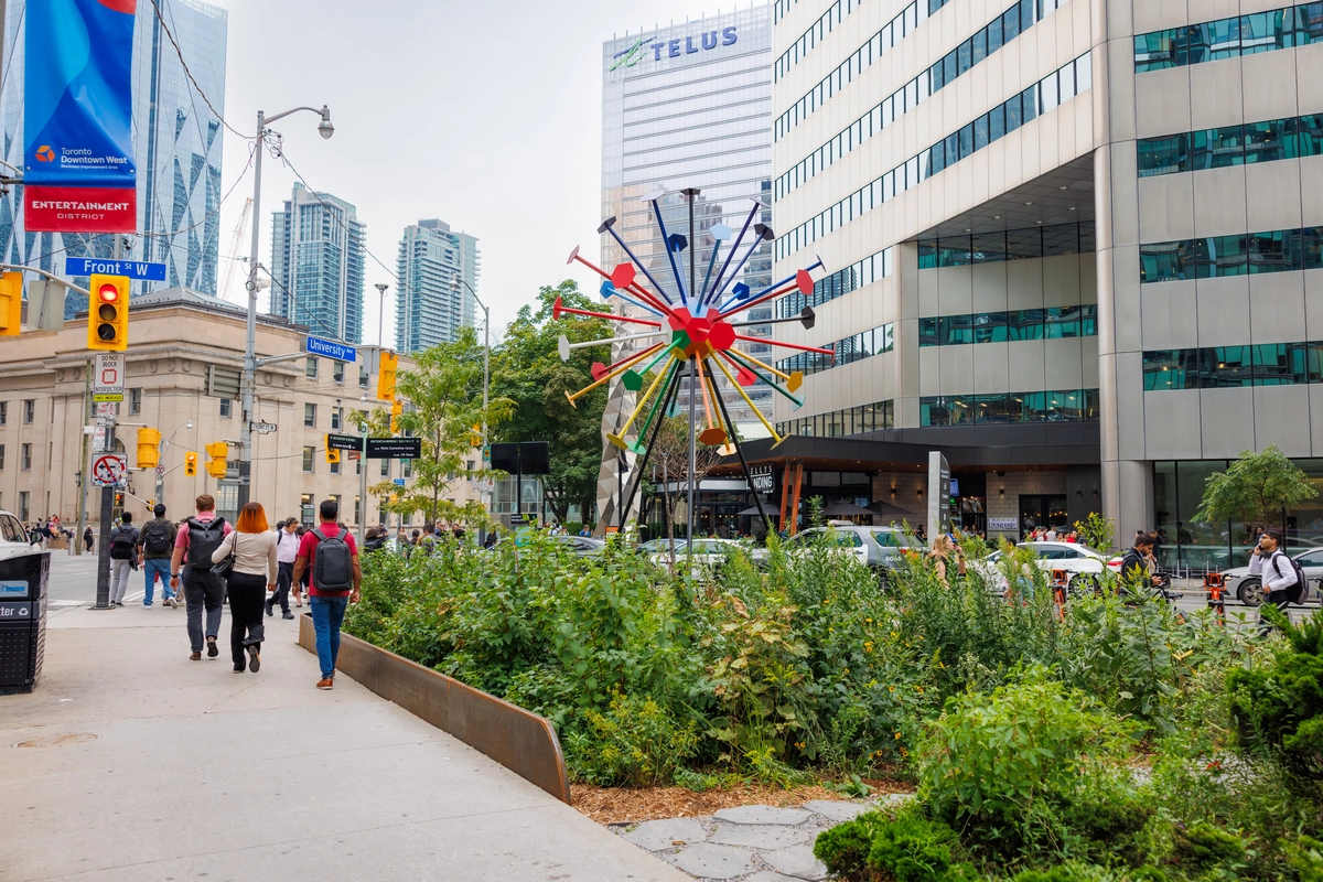 Pedestrians walk on a downtown street corner near a colorful, sunburst-shaped sculpture.