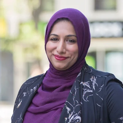 A smiling woman wearing a purple hijab and a patterned dark jacket.