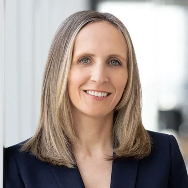 A professional headshot of a smiling woman with shoulder-length hair wearing a dark blazer.